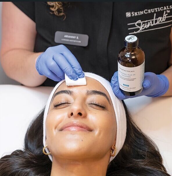 Esthetician in blue gloves applying a chemical peel solution to a smiling client's forehead with a cotton pad