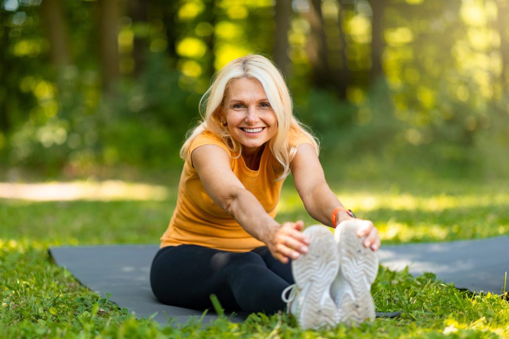 Smiling older woman sitting on a yoga mat in a sunlit park, reaching toward her toes